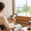旅先の窓辺で電子書籍リーダーを読む女性とキャリーケース