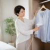 クローゼットで淡いブルーのブラウスを衣類スチーマーで整える女性