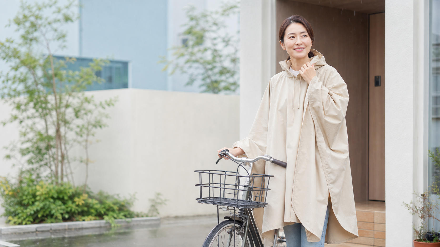 雨の日の玄関でベージュのレインポンチョを着て自転車のそばに立つ女性