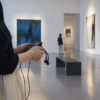 静かな美術館の展示室で抽象画を前に音声ガイド端末とイヤホンを手にする様子