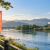 朝の光が差す川辺に無地の旗が並び、山並みと文化施設が遠くに見える秋の風景