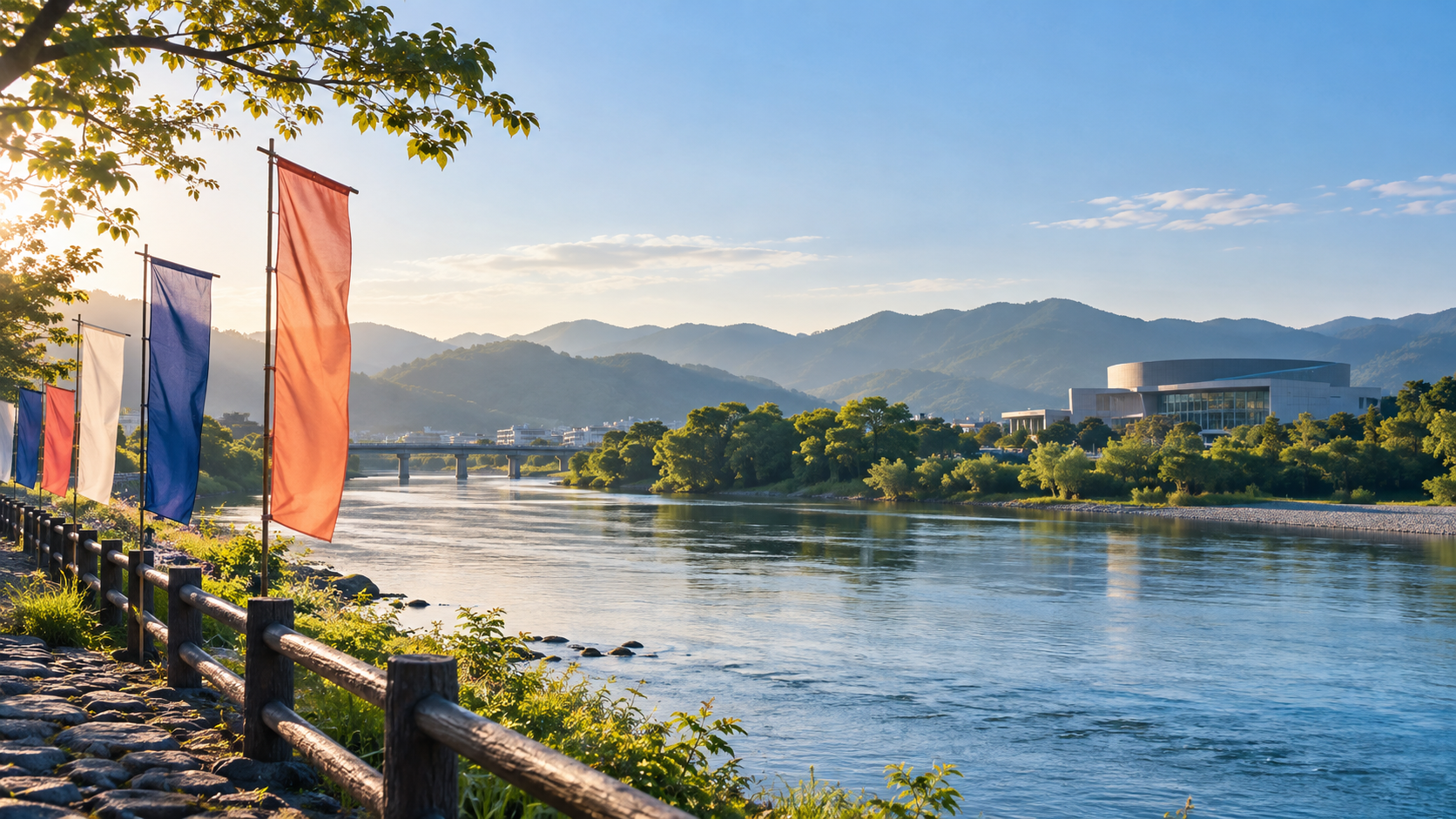 朝の光が差す川辺に無地の旗が並び、山並みと文化施設が遠くに見える秋の風景